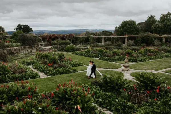 Couple walks hand in hand through a lush garden, featuring vibrant flowers, manicured hedges, and elegant stone pathways.
