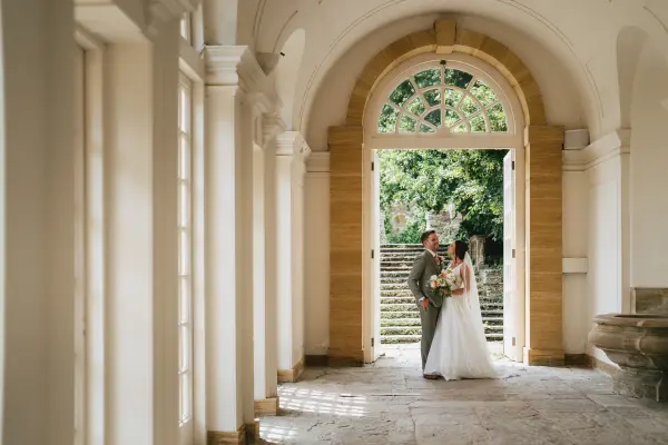 Couple standing in a sunny hallway with arched doorways and stone flooring, framed by lush greenery in the background.