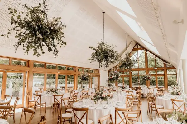 Bright, spacious reception area featuring wooden chairs, elegant round tables, and lush greenery hanging from the ceiling. Perfect for intimate weddings.