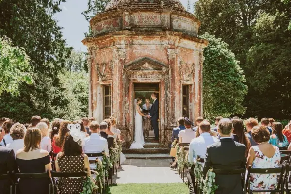 A romantic outdoor ceremony with guests seated in front of a vintage stone structure, surrounded by lush greenery and natural light.