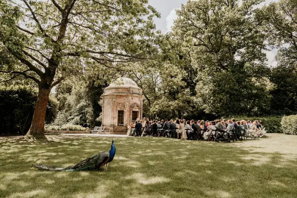 A serene outdoor ceremony space featuring an elegant stone gazebo, surrounded by lush greenery and guests seated on the lawn.