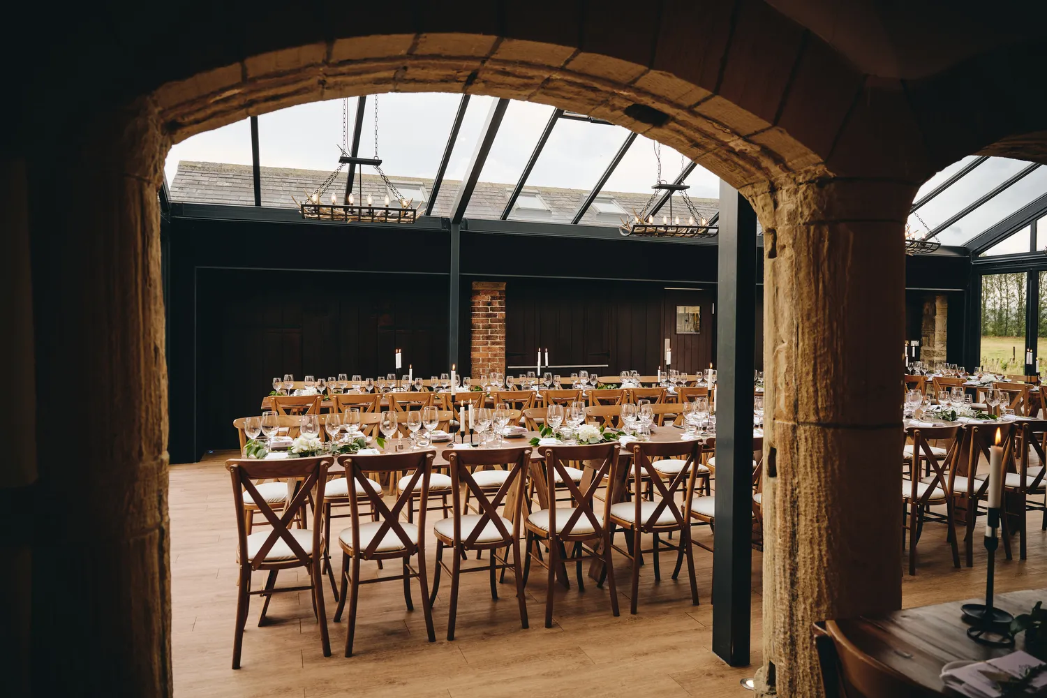 Rustic indoor reception space with wooden tables and cross-back chairs, featuring large glass windows and warm wooden accents.