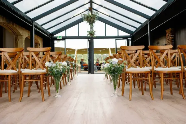Bright and airy indoor ceremony space featuring wooden chairs lined with floral arrangements, set beneath a glass ceiling for a romantic ambiance.