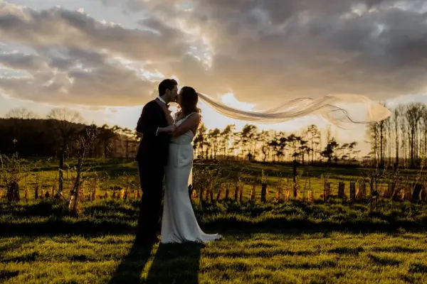A couple shares a romantic kiss as the bride's veil flows gracefully in the golden light of sunset over a picturesque vineyard.