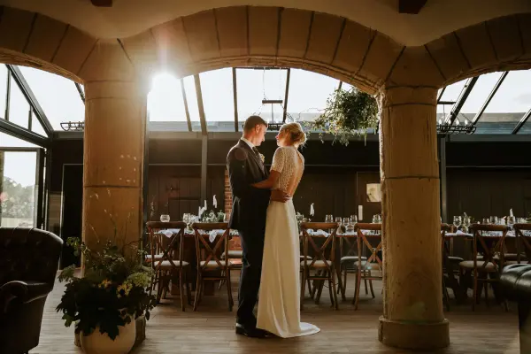 Couple sharing a romantic moment in a sunlit reception area with rustic columns and elegant wooden tables set for a wedding.
