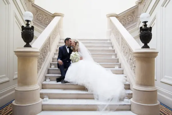 Couple sharing a romantic kiss on an elegant marble staircase, highlighted by ornate railings and soft, ambient lighting.