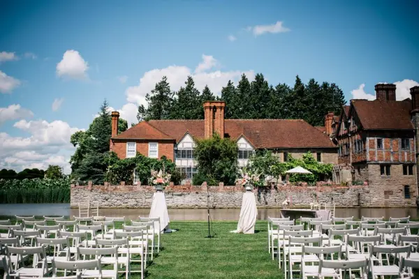 Charming outdoor ceremony space by a serene lake, surrounded by lush greenery and an elegant historic manor backdrop. White chairs arranged for guests.