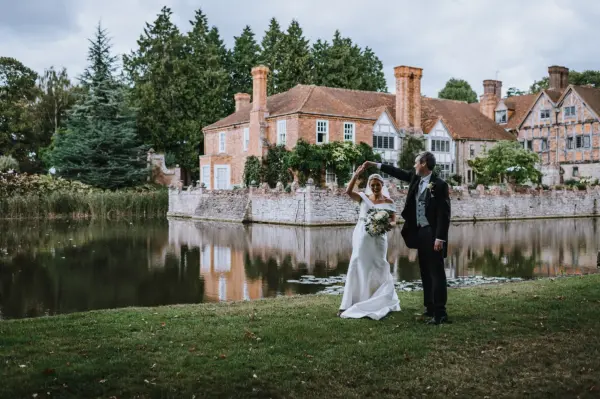 A couple dances by a serene pond, framed by a charming historic estate and lush greenery, creating a romantic wedding backdrop.