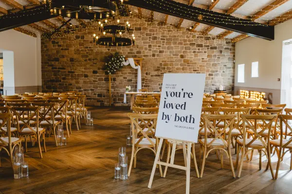 Rustic wedding venue featuring a stone wall, wooden beams, elegant chandeliers, and charming cross-back chairs arranged for a ceremony.