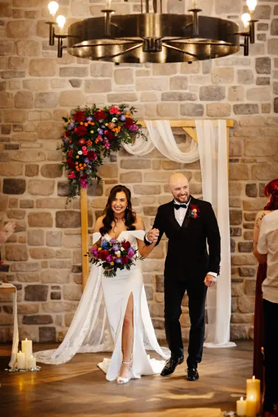 Elegant indoor ceremony with stone walls, floral arch, soft drapery, candlelight, and a couple celebrating their union.