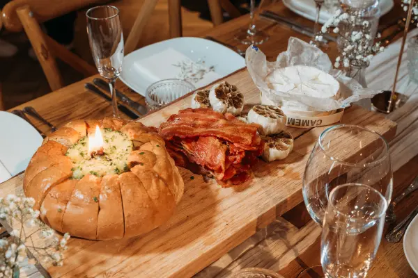 A rustic wooden table set for a wedding featuring gourmet bread, crispy bacon, roasted garlic, and elegant glassware, surrounded by delicate flowers.