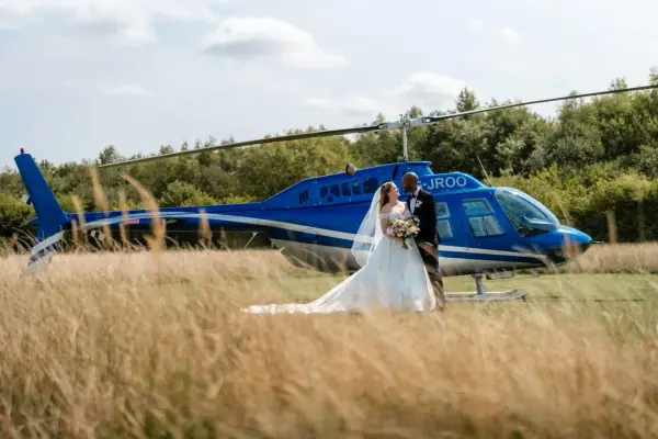 Couple embracing in a golden field, with a blue helicopter parked nearby, creating a unique backdrop for a memorable wedding celebration.