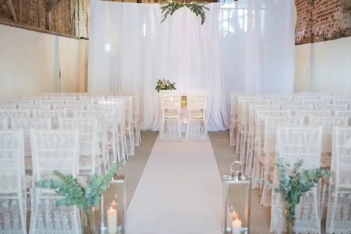 Charming rustic barn venue with elegant white drapery, clear acrylic chairs, and a romantic aisle adorned with candles and greenery.