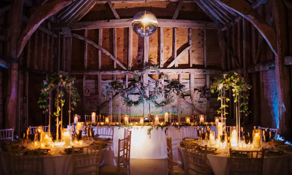 Rustic barn interior featuring elegant tables adorned with candles and greenery, highlighted by warm lighting and a disco ball overhead.