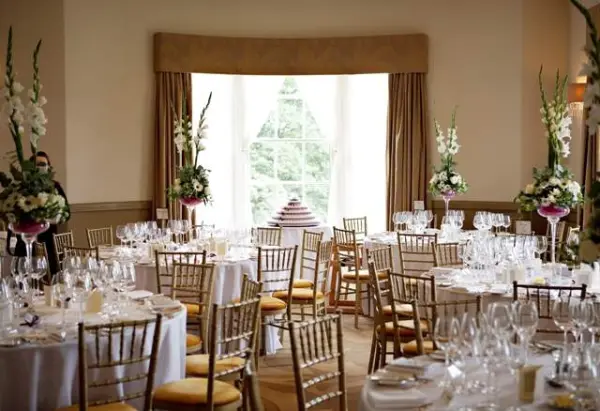 Elegant reception space featuring round tables set with fine china, crystal glassware, and tall floral centerpieces, alongside a large window view.