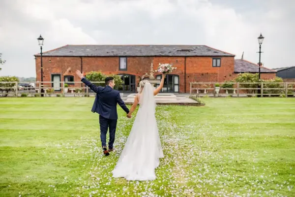 Bride and groom walking hand in hand on a flower-strewn path, with a rustic red brick venue and lush green lawn in the background.