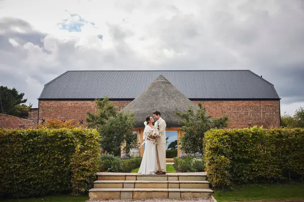 A couple embraces on steps in front of a rustic brick barn, framed by lush hedges and an overcast sky, perfect for a romantic ceremony.