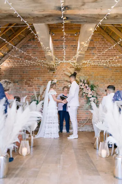 Charming rustic ceremony space with exposed brick walls, string lights, and elegant floral arrangements, framed by soft pampas grass decor.
