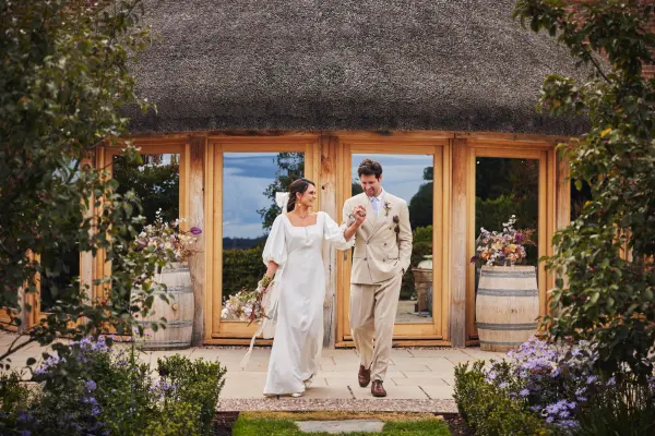 Couple walking hand-in-hand towards a charming thatched-roof venue surrounded by lush gardens and seasonal blooms.