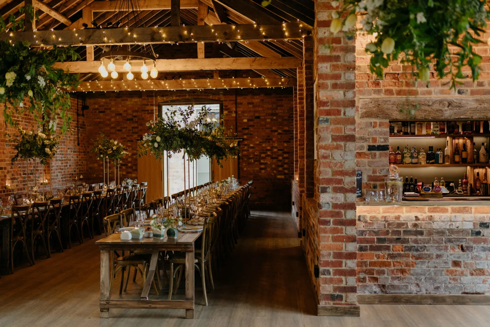 Rustic barn venue featuring exposed brick walls, elegant wooden tables, twinkling string lights, and lush greenery for a charming atmosphere.