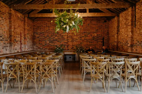 Rustic ceremony space featuring warm exposed brick walls, wooden cross-back chairs, and a lush greenery chandelier overhead.