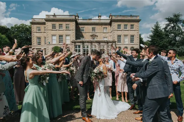 A joyful wedding celebration outside a grand estate, with guests throwing petals around the happy couple in elegant attire.