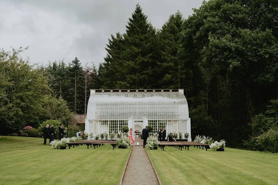 Lush green lawn with a charming glass greenhouse, beautifully framed by trees, perfect for an intimate outdoor wedding ceremony.