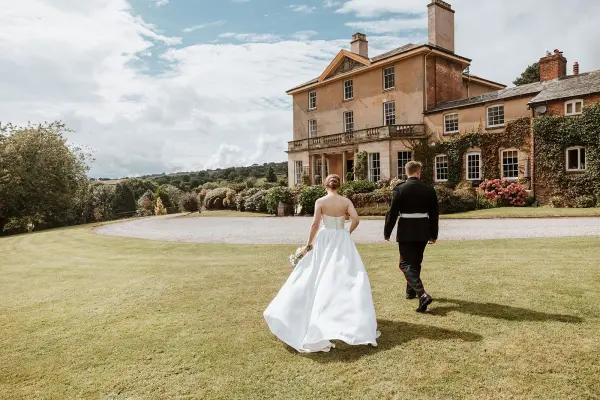 A couple walks hand-in-hand across lush green lawns towards a charming, historic estate adorned with climbing greenery and blooming flowers.