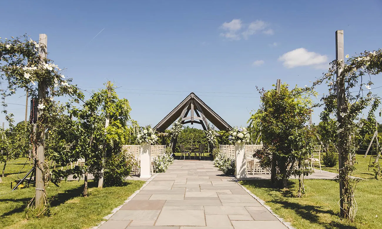 Charming outdoor ceremony space featuring a stone pathway framed by lush greenery and elegant floral arrangements, leading to a striking pavilion.