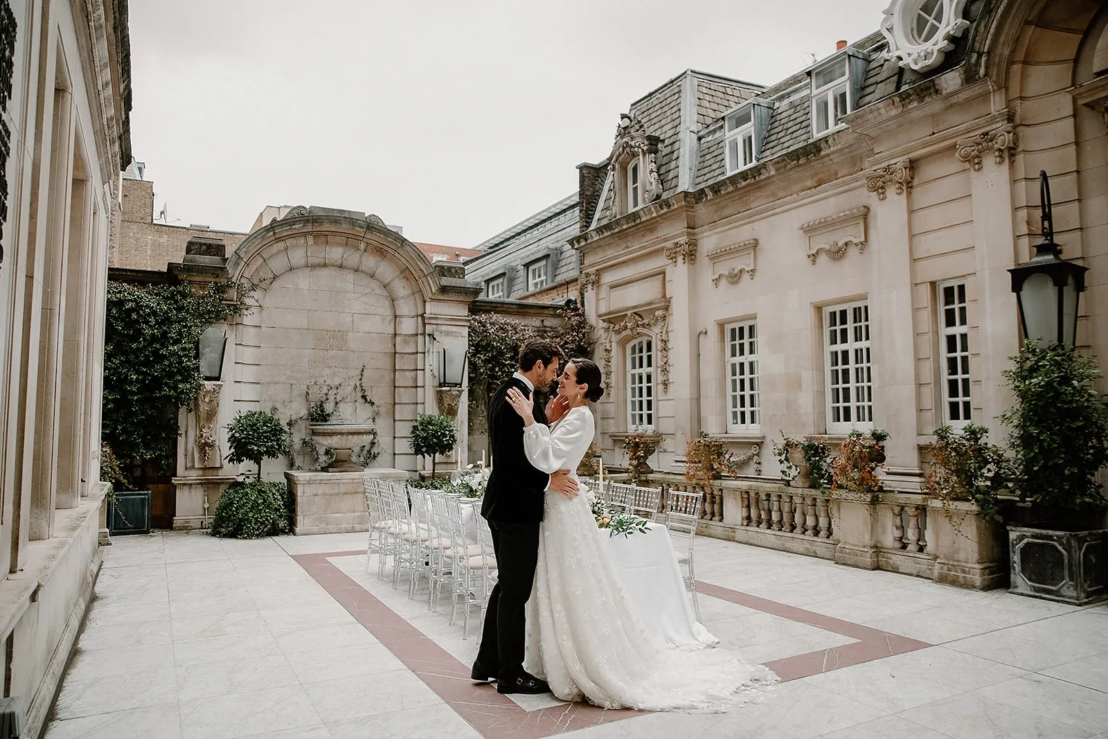 Intimate outdoor wedding ceremony on a stone terrace, featuring elegant architecture, greenery, and a charming fountain backdrop.