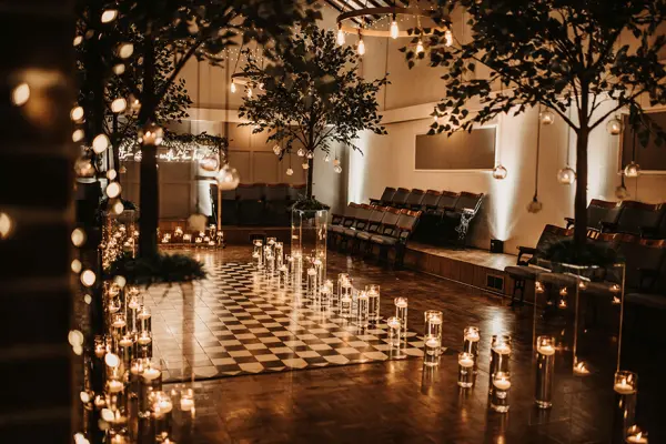 Intimate indoor ceremony space featuring checkered flooring, soft lighting, hanging globes, and elegant trees surrounded by glowing candles.