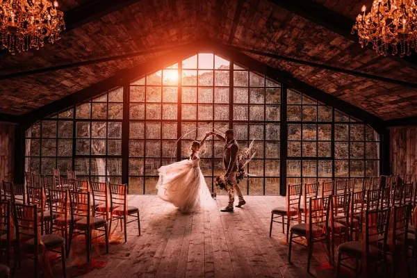 Couple dancing in a rustic venue with large industrial windows, warm lighting, and chandeliers, surrounded by elegant wooden chairs.