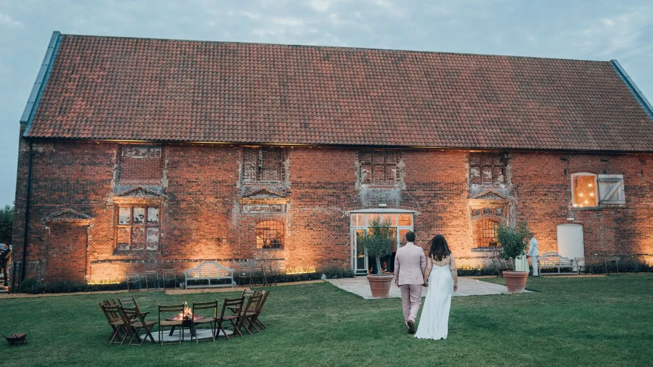 A couple walks hand-in-hand towards a beautifully lit rustic barn, surrounded by a green lawn and cozy outdoor seating.
