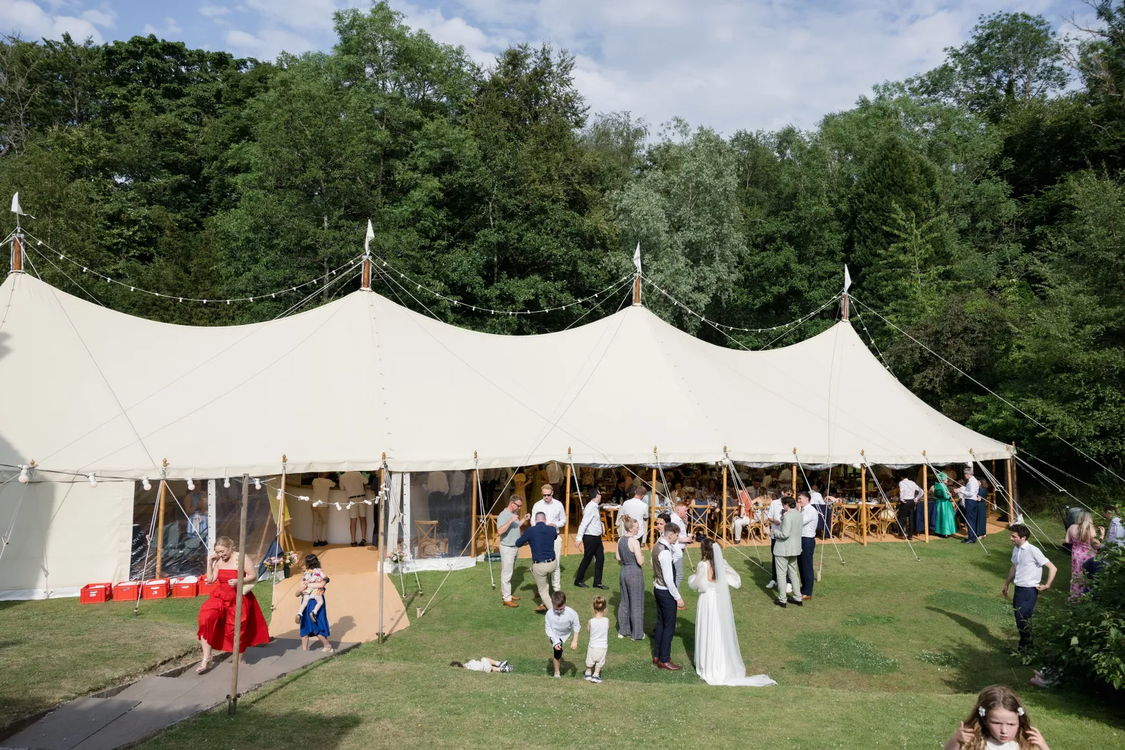 An elegant outdoor wedding tent set in lush greenery, featuring guests mingling and enjoying a sunny day celebration.