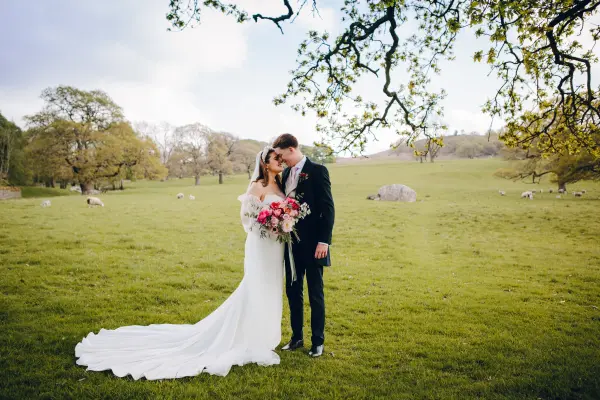 A romantic couple shares a kiss in a lush, green field, surrounded by trees and grazing sheep, perfect for an outdoor wedding setting.