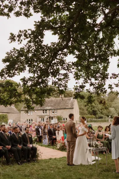 Intimate outdoor wedding ceremony under a large oak tree, with guests seated on lush grass and a charming rustic venue in the background.