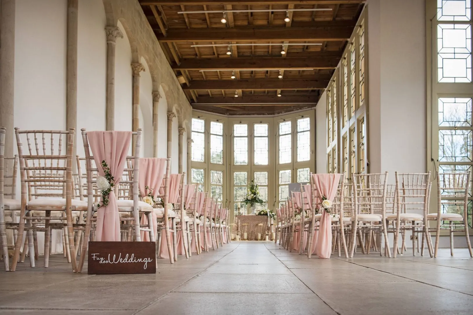 Bright and airy indoor ceremony space featuring elegant chiavari chairs, blush draping, and large windows for a romantic ambiance.