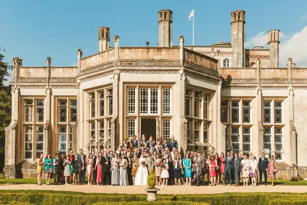 A large group gathered in front of a grand, historic mansion with tall windows and elegant architectural details, surrounded by lush greenery.