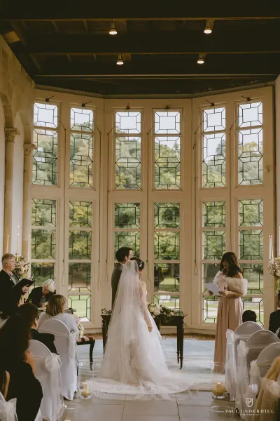 Elegant indoor ceremony featuring a couple exchanging vows beneath stunning stained glass windows, surrounded by seated guests.