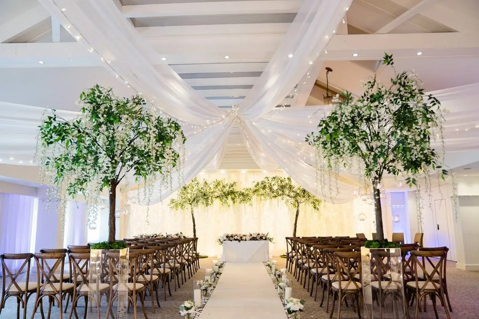 Elegant wedding ceremony space with draped fabric overhead, greenery, and cross-back chairs lining the aisle for an enchanting atmosphere.