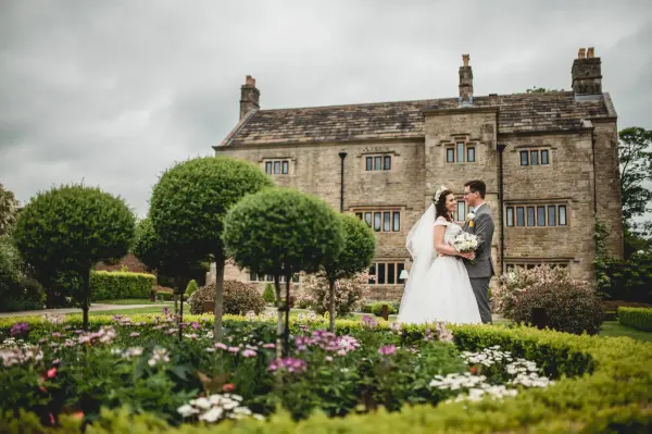 Couple sharing a moment in front of a charming stone mansion, surrounded by lush gardens and vibrant flower beds.