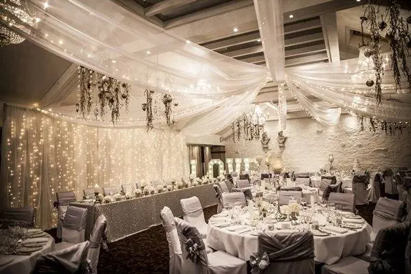 Elegant wedding reception featuring sparkling fairy lights, draped fabric, and stylish tables dressed in silver and white decor.