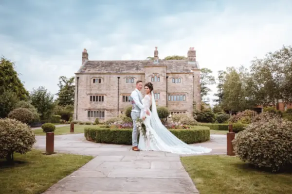 A bride and groom pose on a stone pathway surrounded by manicured gardens and a charming historic mansion in the background.