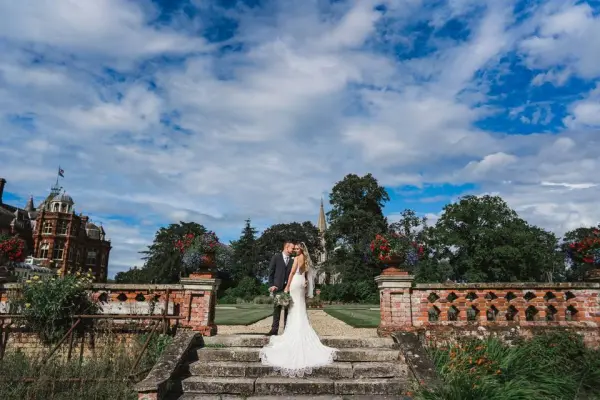 A couple shares a romantic kiss on stone steps, framed by lush gardens and a historic manor under a dramatic sky.