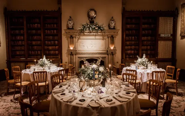 Intimate dining area featuring elegant round tables set with fine china, surrounded by bookshelves and a cozy fireplace backdrop.