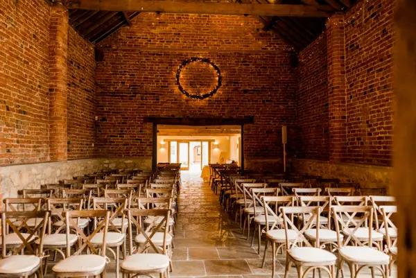 Rustic indoor ceremony space featuring exposed brick walls, wooden beams, and rows of elegant wooden chairs leading to bright double doors.