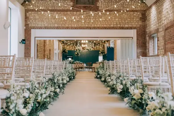 Elegant ceremony space featuring rows of wooden chairs, a floral-lined aisle, and twinkling string lights against rustic stone walls.