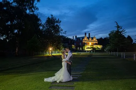 Couple embracing on a stone path with a picturesque mansion illuminated in the background, surrounded by lush greenery at twilight.