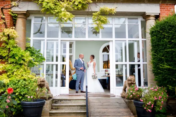 Couple stands at a grand glass entrance, framed by lush greenery and colorful flowers, inviting a charming garden wedding atmosphere.