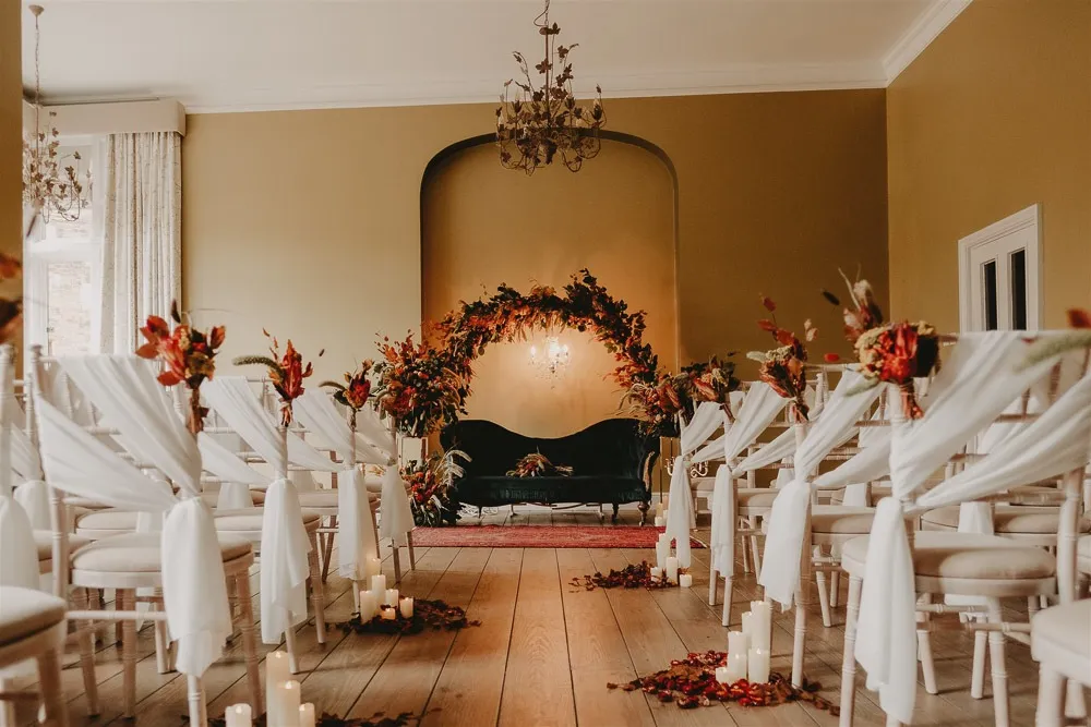 Elegant indoor ceremony space featuring draped chairs, floral arrangements, and a cozy seating area with candles on a wooden floor.